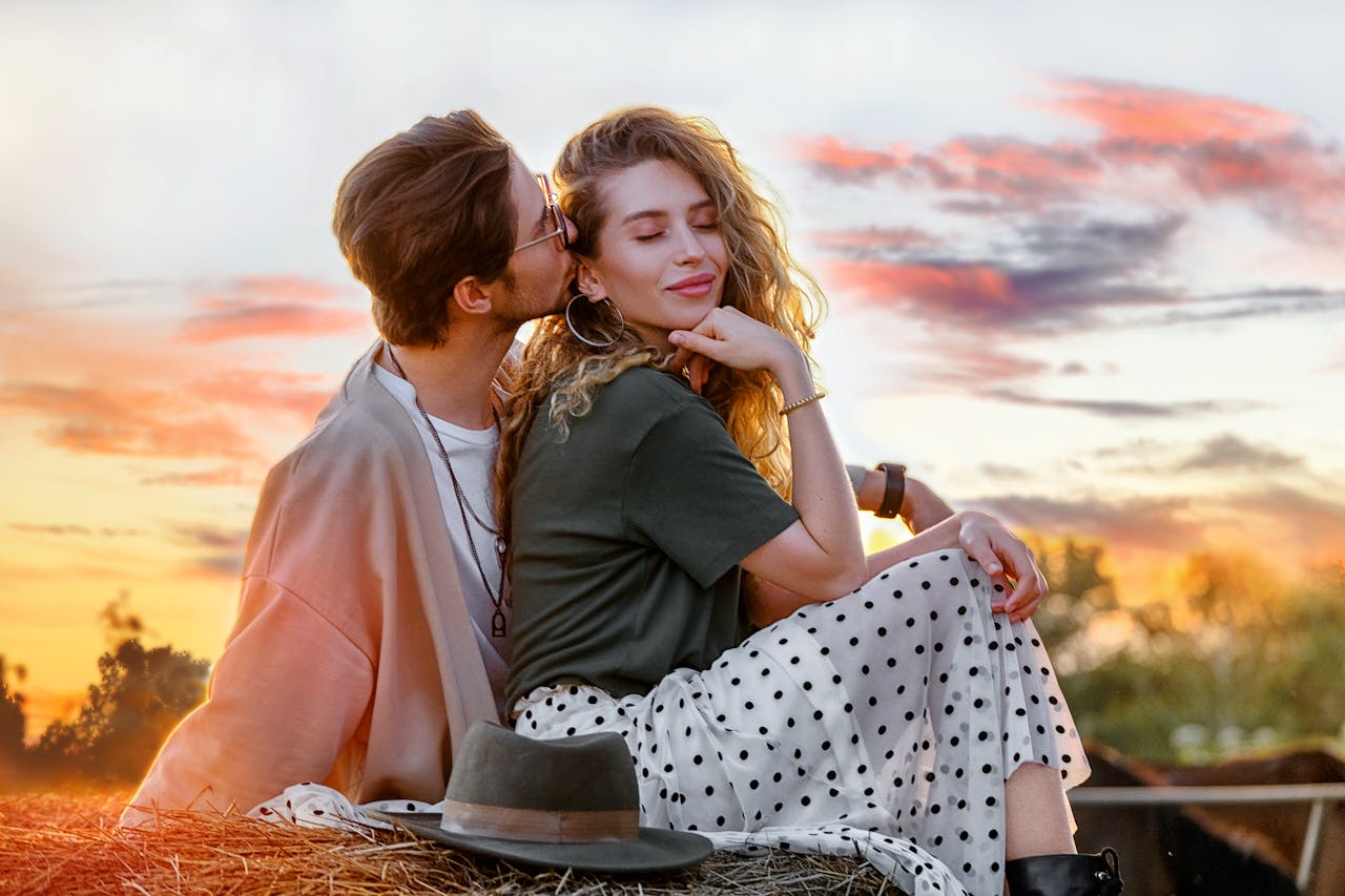 Side view serene young couple in stylish wear kissing and bonding while sitting on haymow against picturesque sunset sky